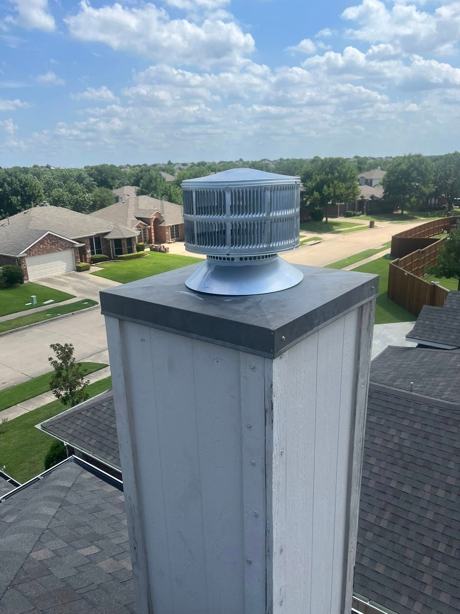 Chimney with a rotating vent cap on a roof, overlooking a residential neighborhood on a sunny day.