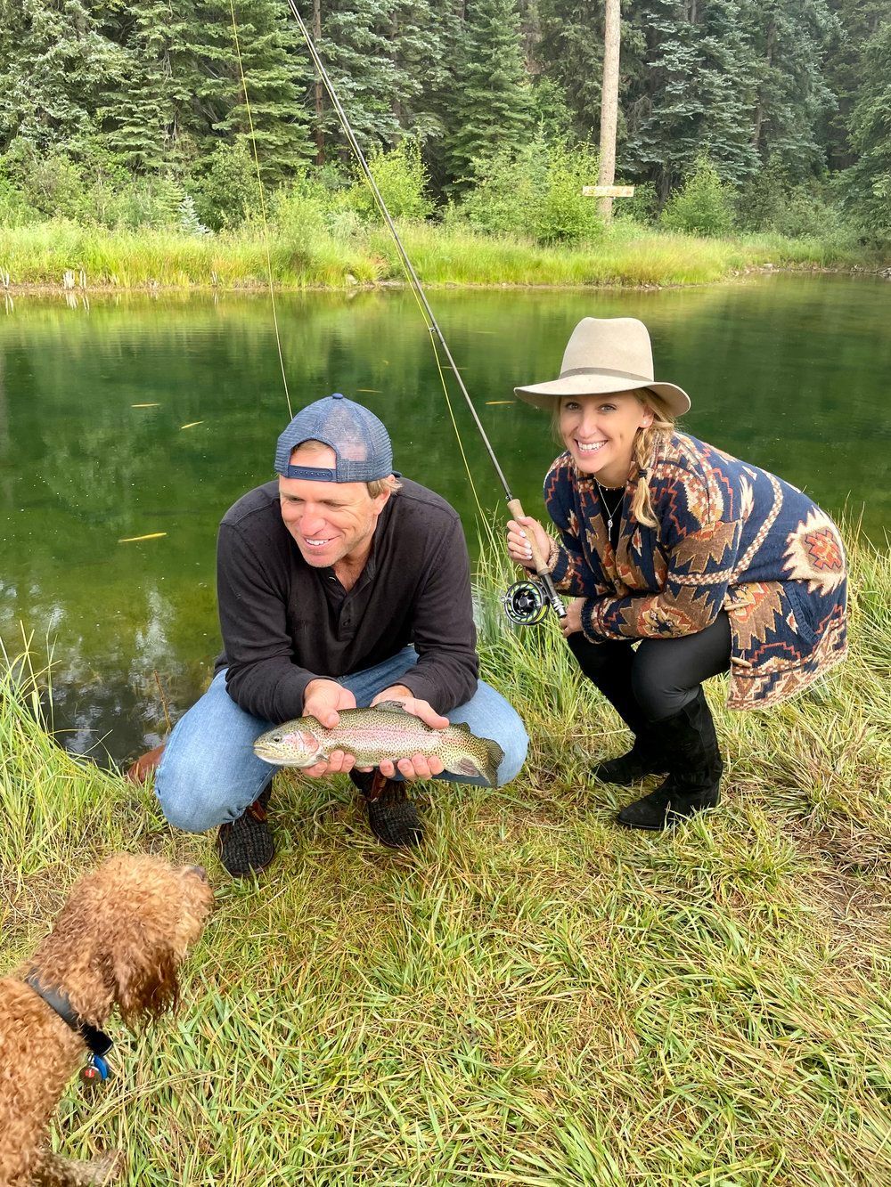 A man and a woman are kneeling next to a lake holding a fish.
