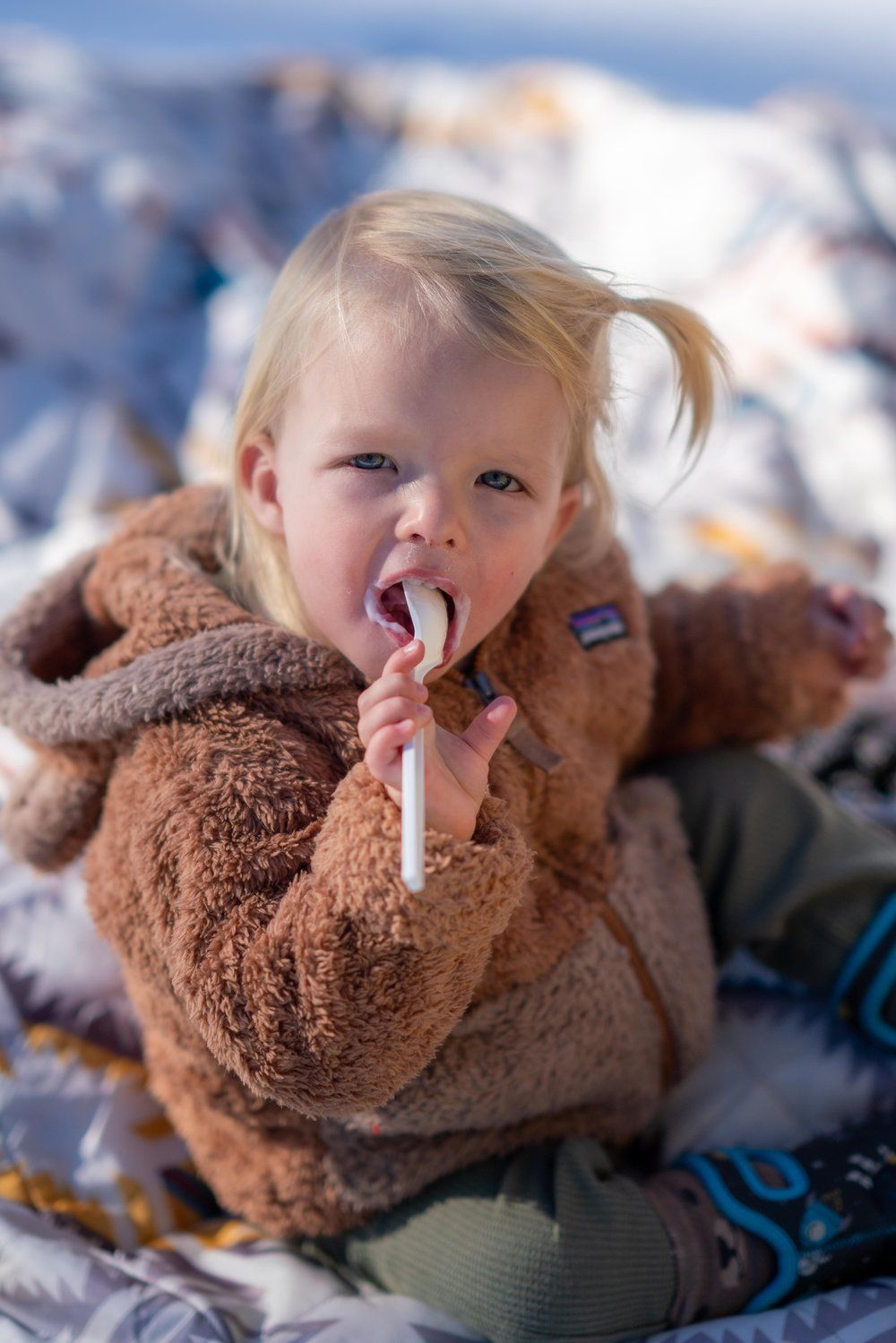 A little girl is sitting on a blanket in the snow eating a marshmallow.