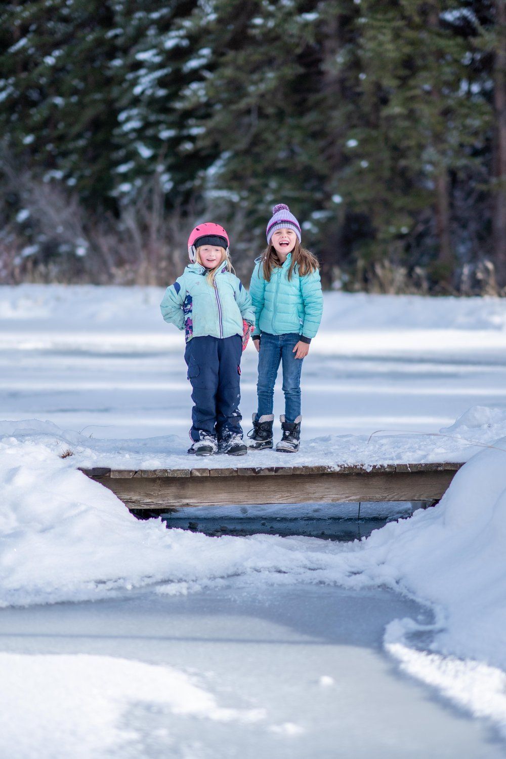 Two little girls are standing on a bridge over a frozen lake.