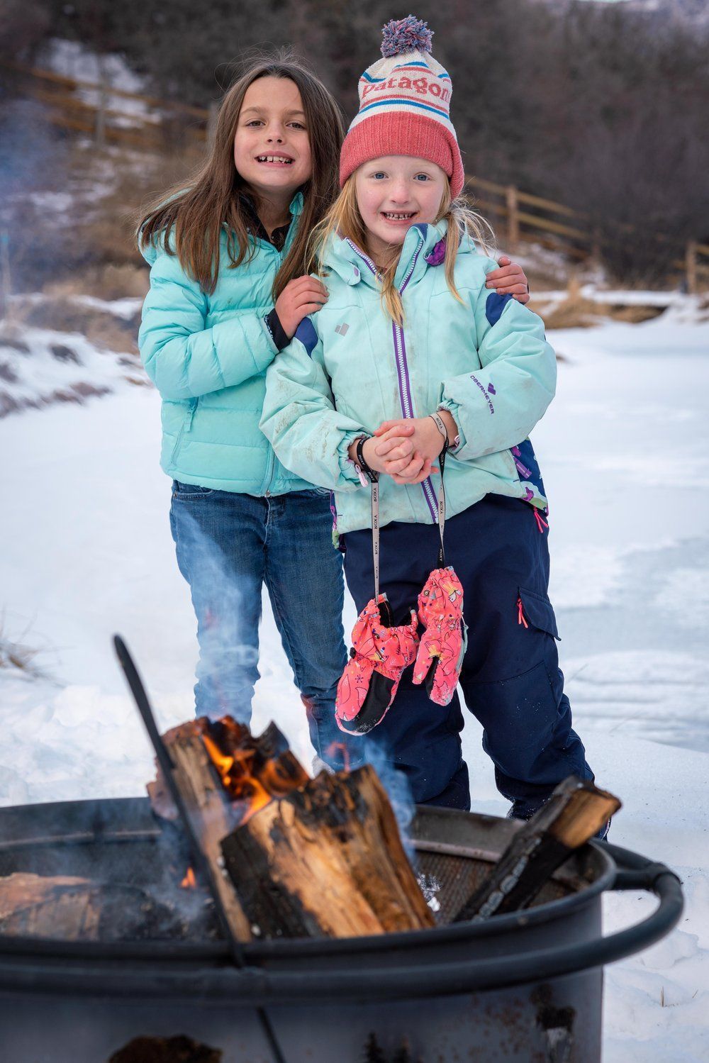 Two young girls are standing next to a fire pit in the snow.