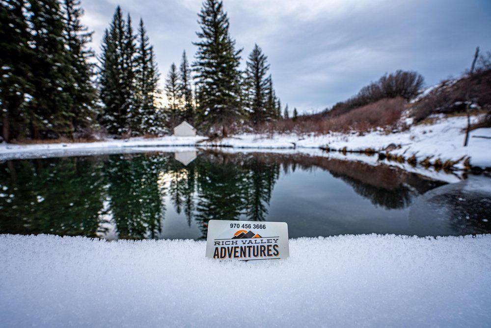 A license plate that says adventures is sitting in front of a snow covered pond.
