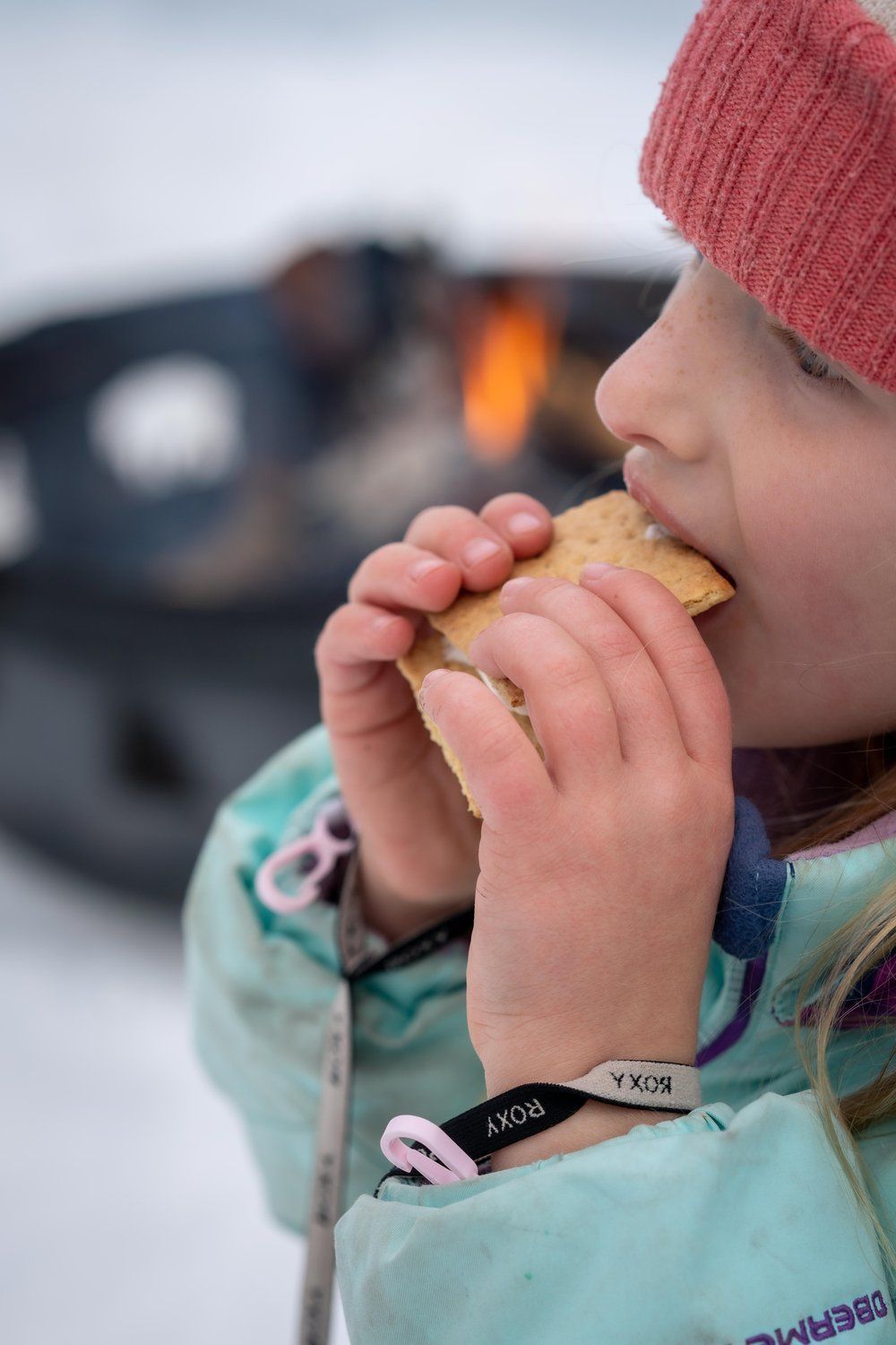 A little girl is eating a s'more in the snow.