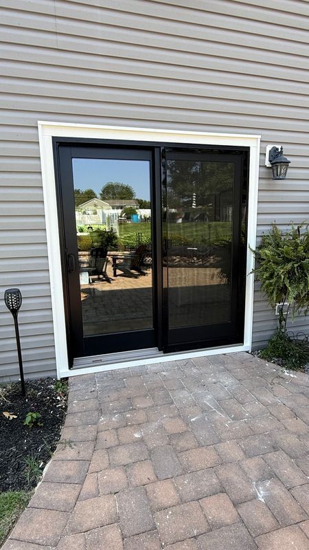 A black-framed sliding glass door with a screen door set into light gray vinyl siding above a stone patio.