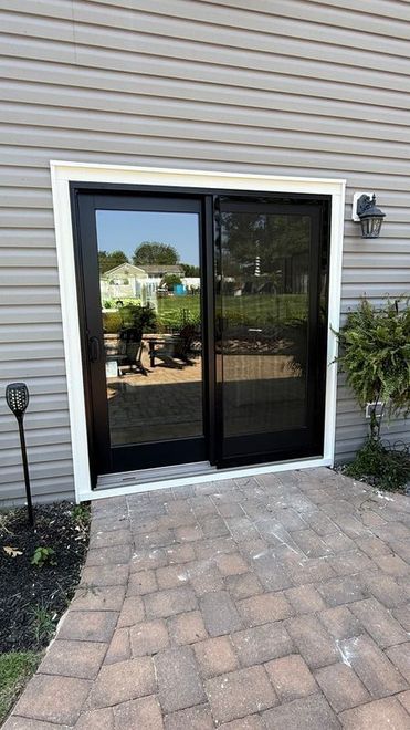 A black-framed sliding glass door with a screen door set into light gray vinyl siding above a stone patio.