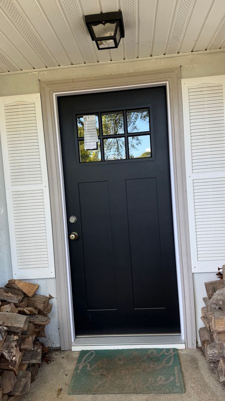 A black front door with a window pane at the top, flanked by white shutters and stacks of firewood on a concrete porch.