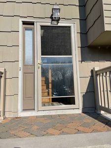 Tan exterior door with a matching screen door and a narrow sidelight window, set on a brick patio.