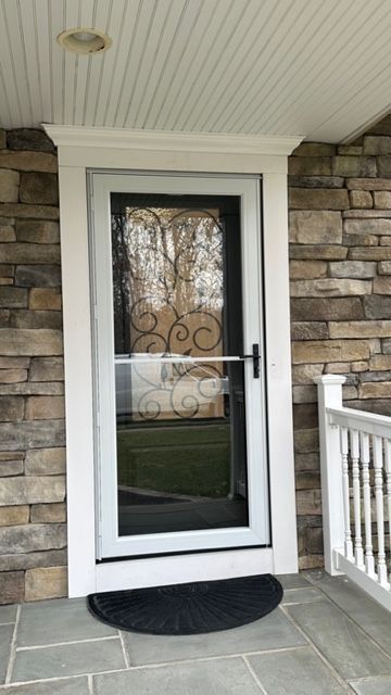 A white storm door with a scrollwork design on the glass, set in a stone-clad exterior doorway with a stone porch floor.