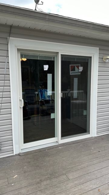 A white-framed sliding glass door installed on the exterior of a home with gray vinyl siding and a wooden deck.
