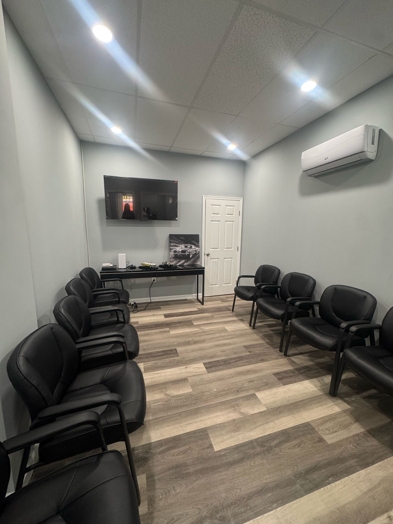 A small waiting room with rows of black chairs, a wall-mounted TV, a table, and a white door under an air conditioner. | Superior Auto Repair and Tires