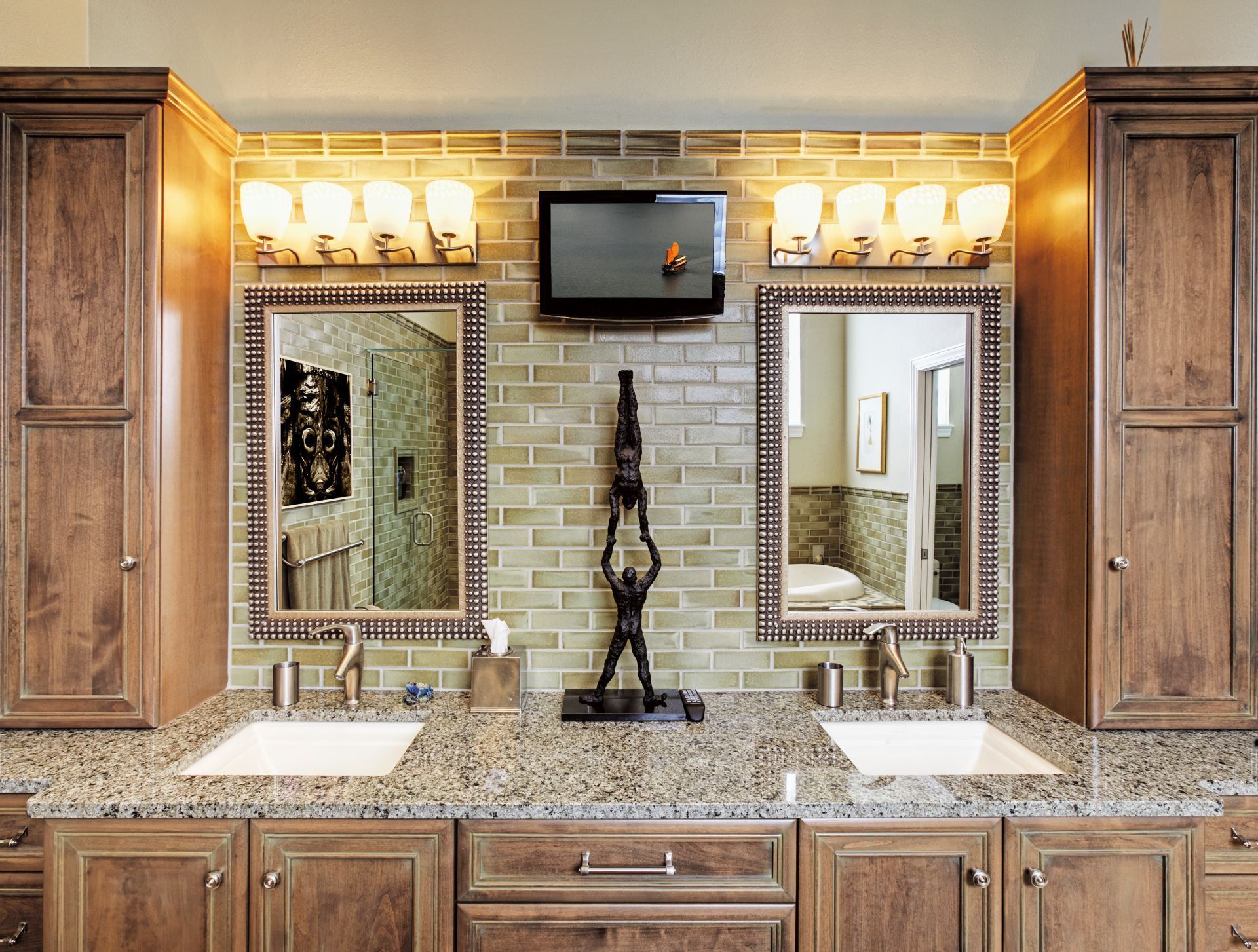 Bathroom vanity with mirrors, lights, and small TV above sinks, framed by wooden cabinets and granite countertop.