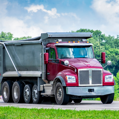 A red dump truck is driving down a road next to a grassy field.