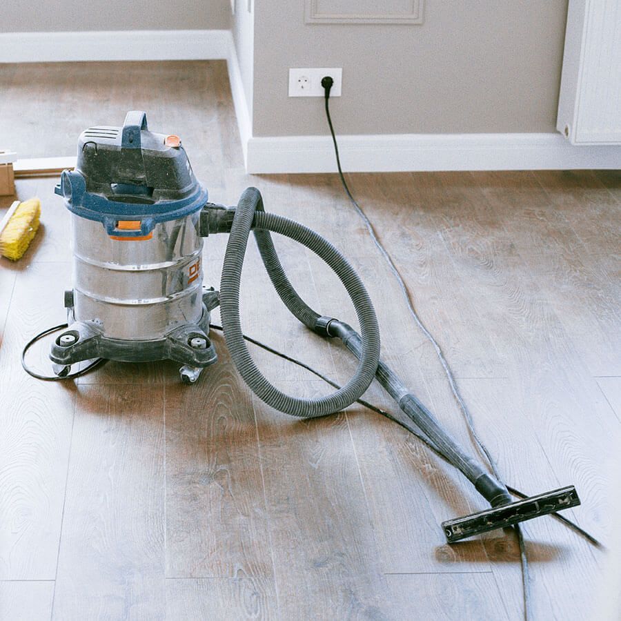 Wet/dry vacuum cleaner on a wooden floor, near a wall with an electrical outlet; cord is plugged in.