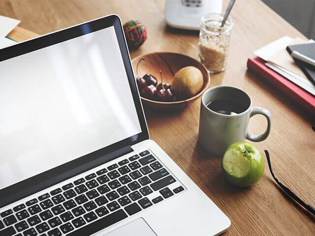 Laptop open on a wooden table with breakfast items: fruit, coffee, and an apple.
