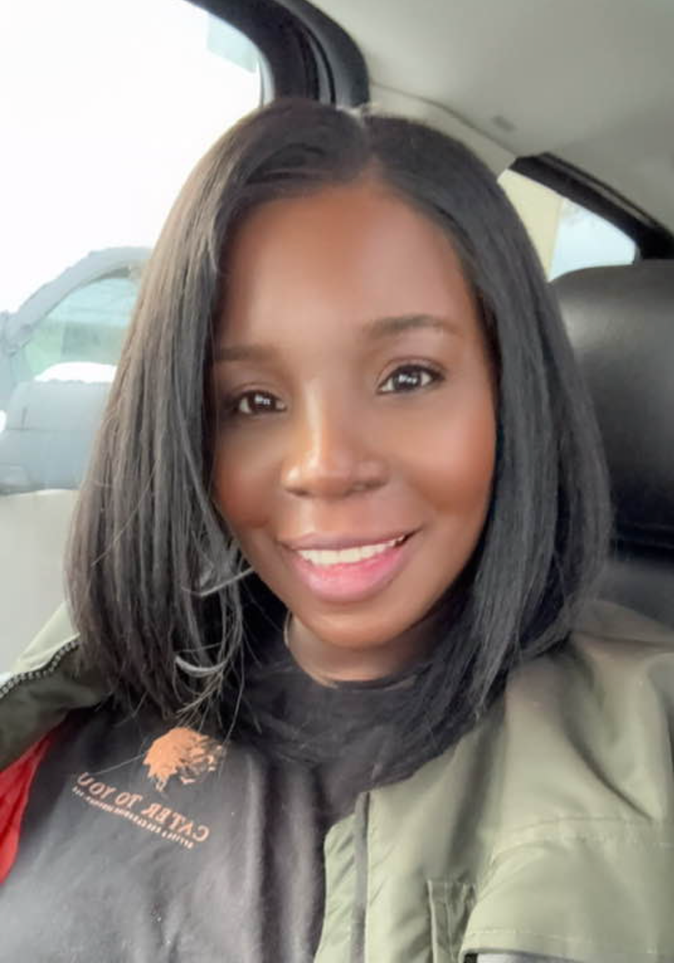 Woman with black hair, smiling, wearing an olive-green jacket and a dark shirt, indoors.