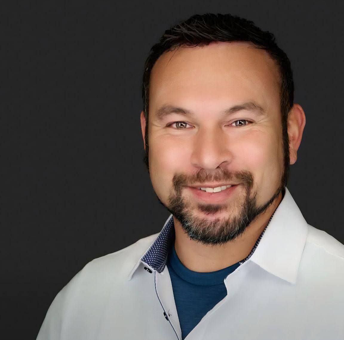 Man with a goatee smiling, wearing a white shirt over a blue shirt, against a gray background.