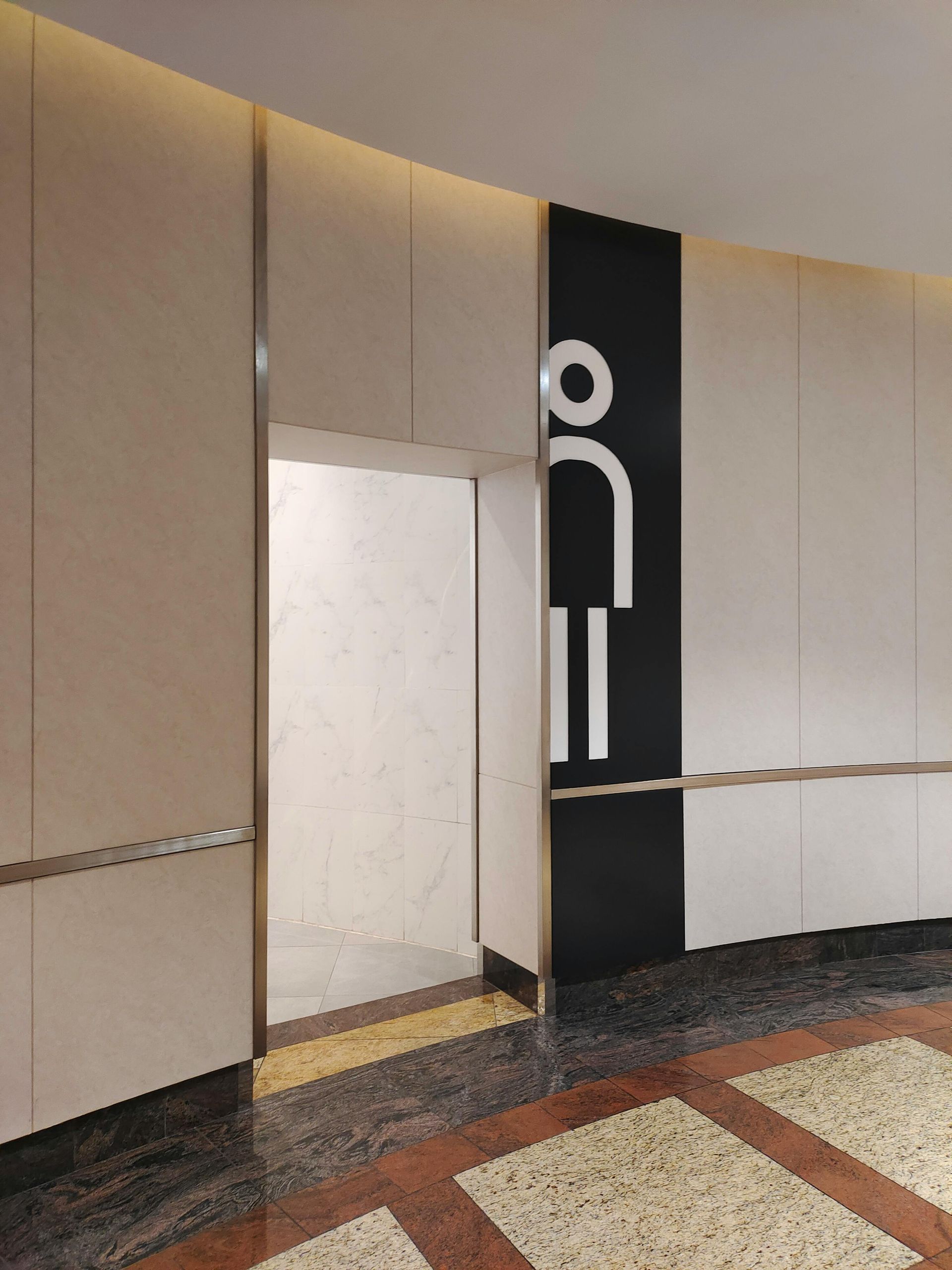 Men's restroom doorway with sign. Beige walls, black sign with white figure, mosaic floor.