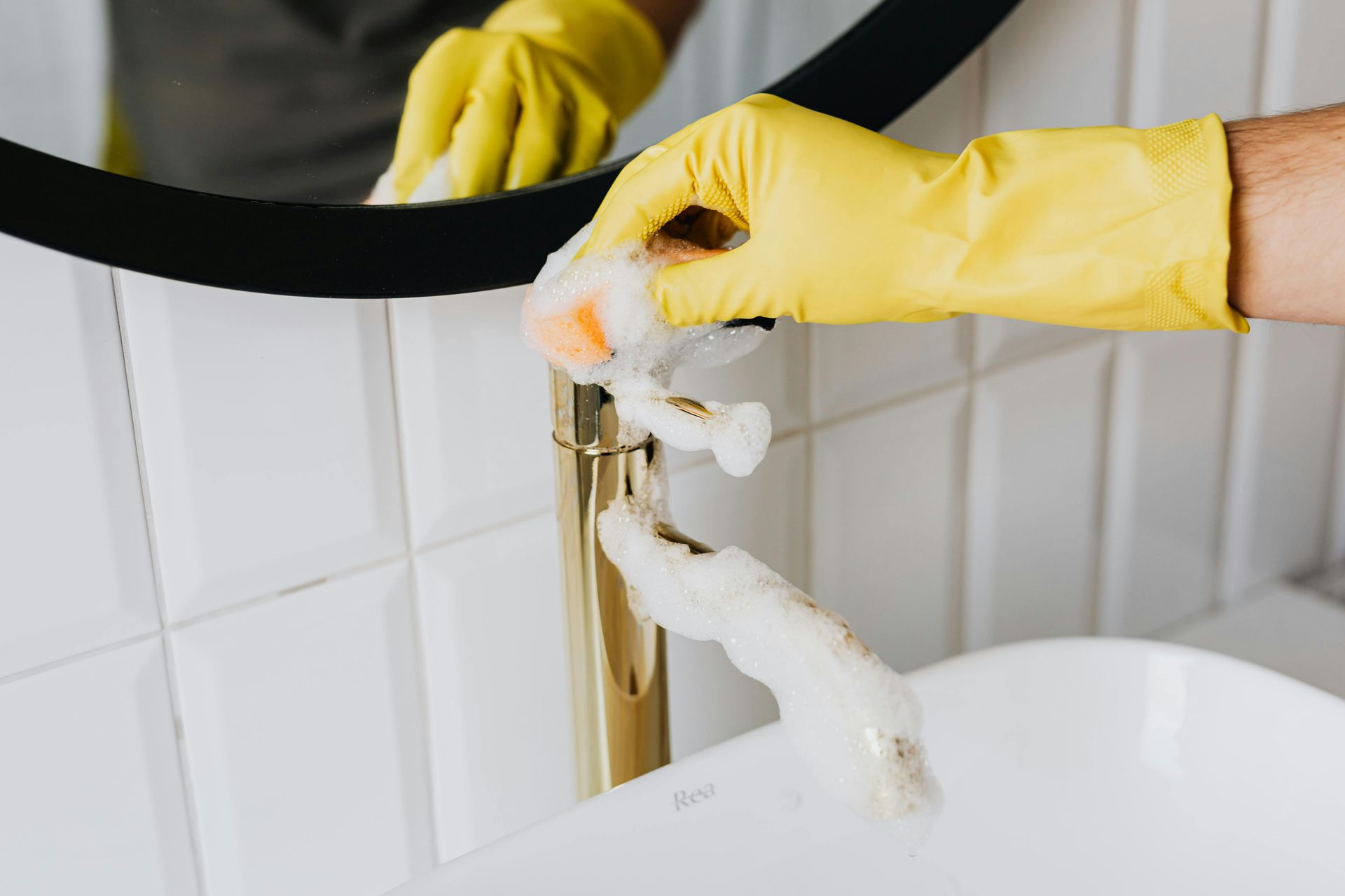 Hand in yellow glove cleaning a gold faucet with soap suds in a white bathroom.