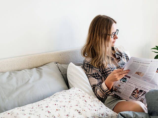 Woman in glasses reading newspaper in bed.