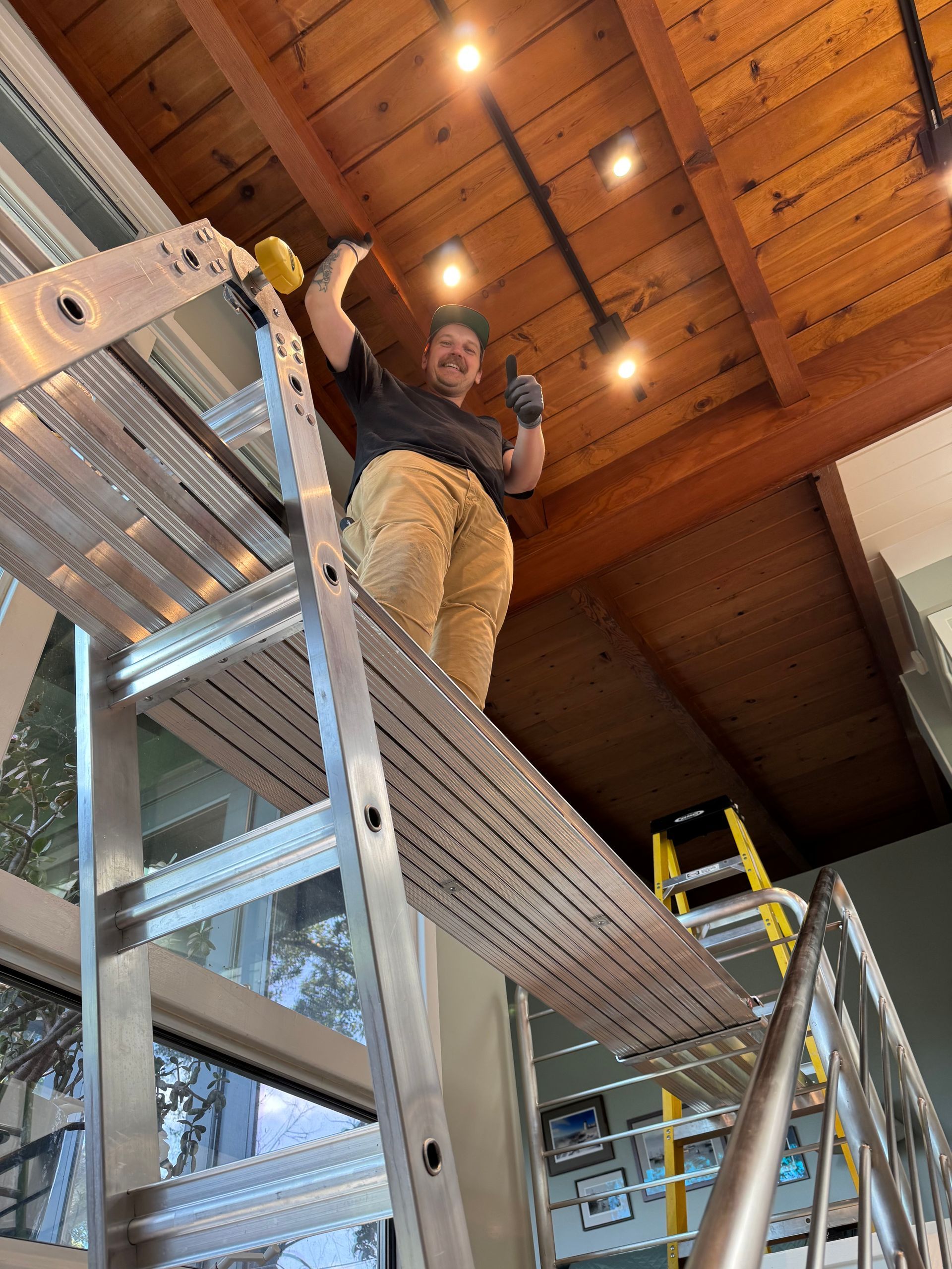Man on a metal scaffold giving a thumbs up, working on a wood-paneled ceiling with track lighting.