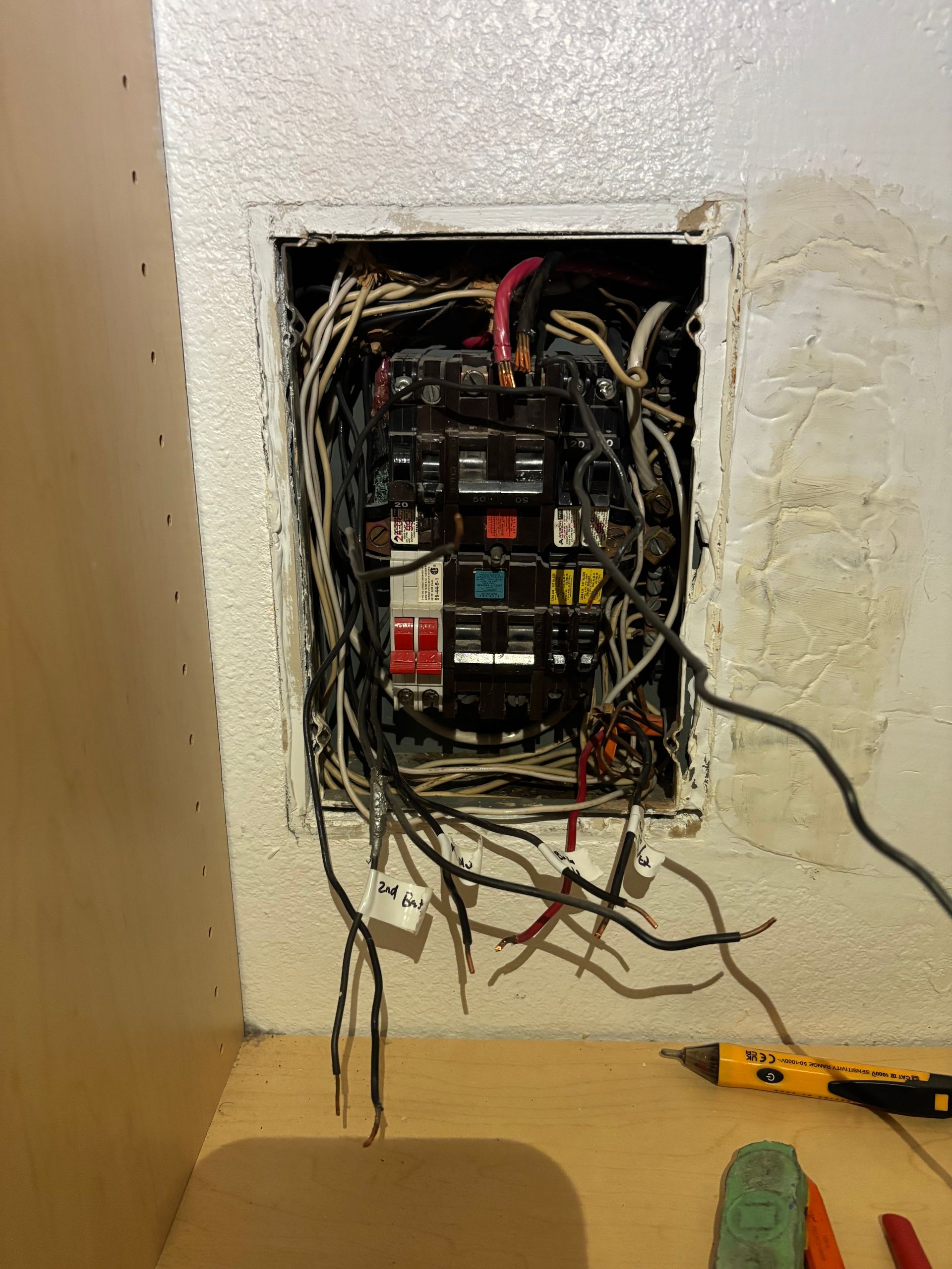 Electrical panel in a wall. Numerous wires, breakers, and a partially open box.