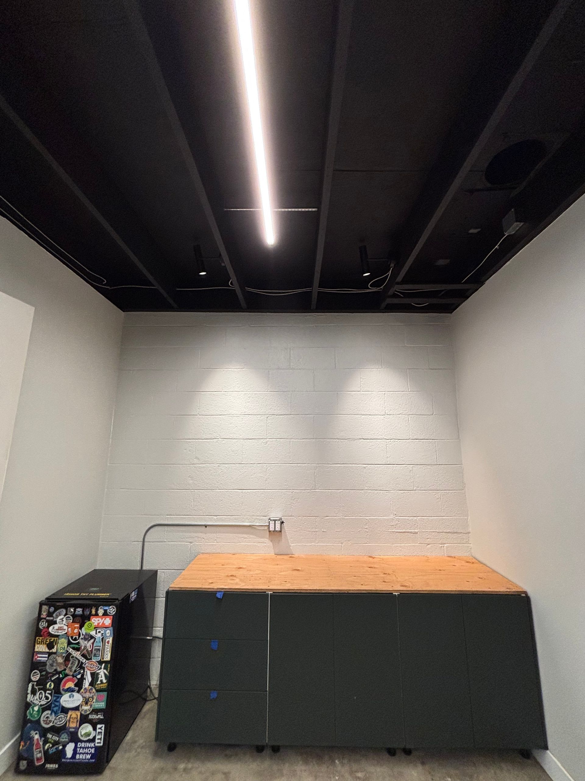 Workshop with black ceiling, white walls, and a wooden workbench. A black cabinet with stickers is on the left.