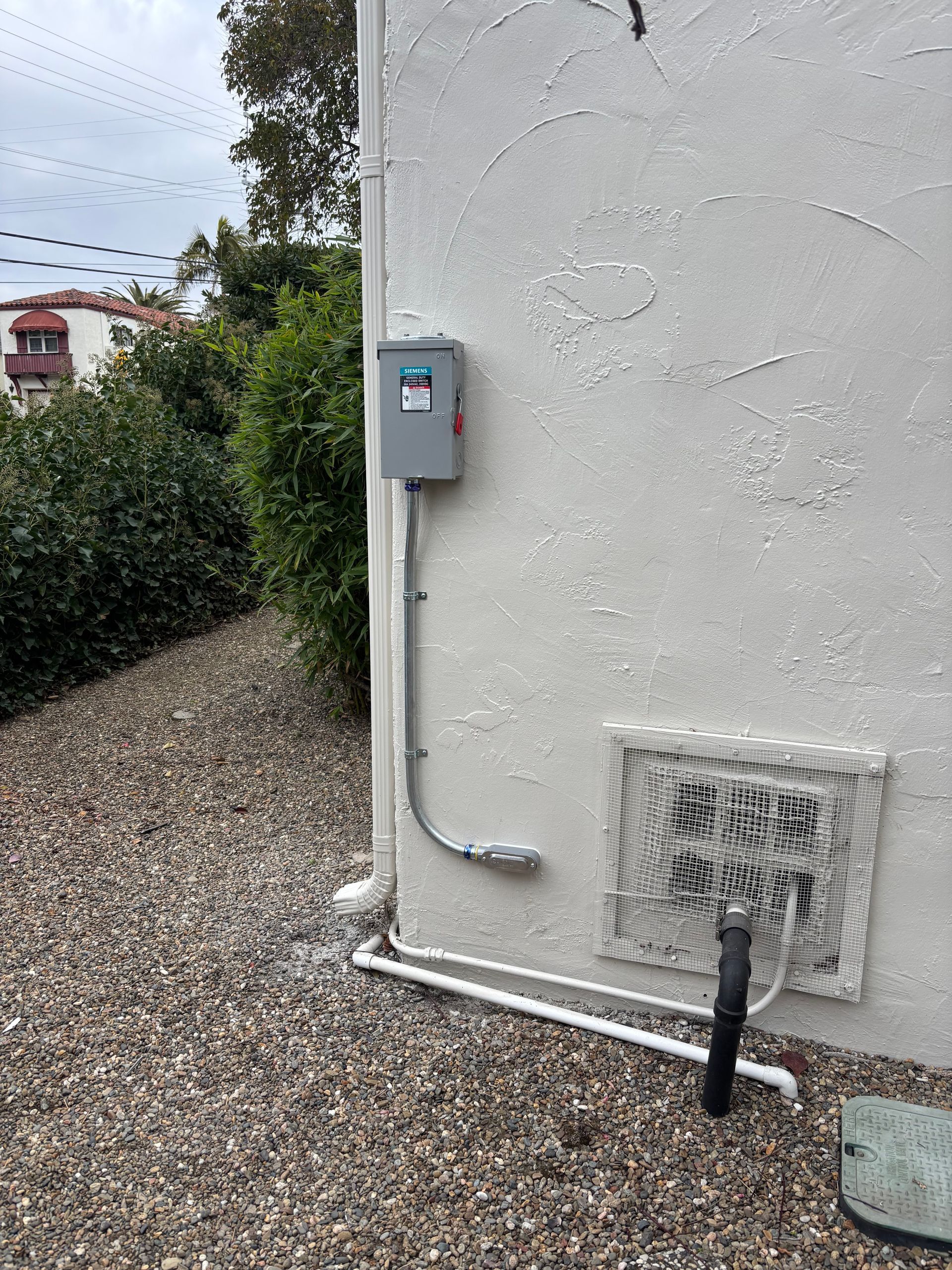 Gray electrical box mounted on a textured white wall near a gravel area, greenery, and pipes.