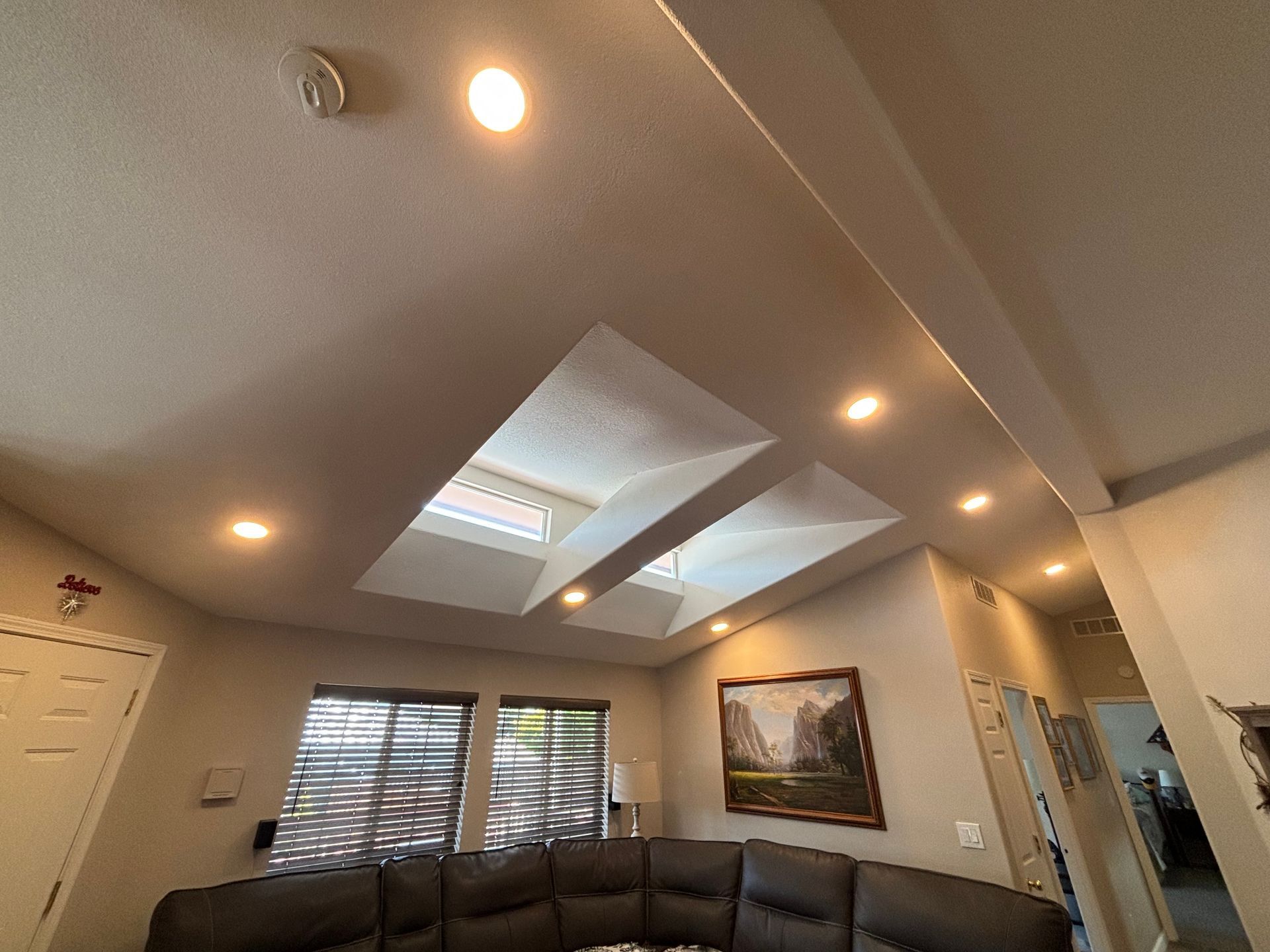 Interior view of a living room with skylights and recessed lighting. Dark sectional sofa, artwork, and window blinds.