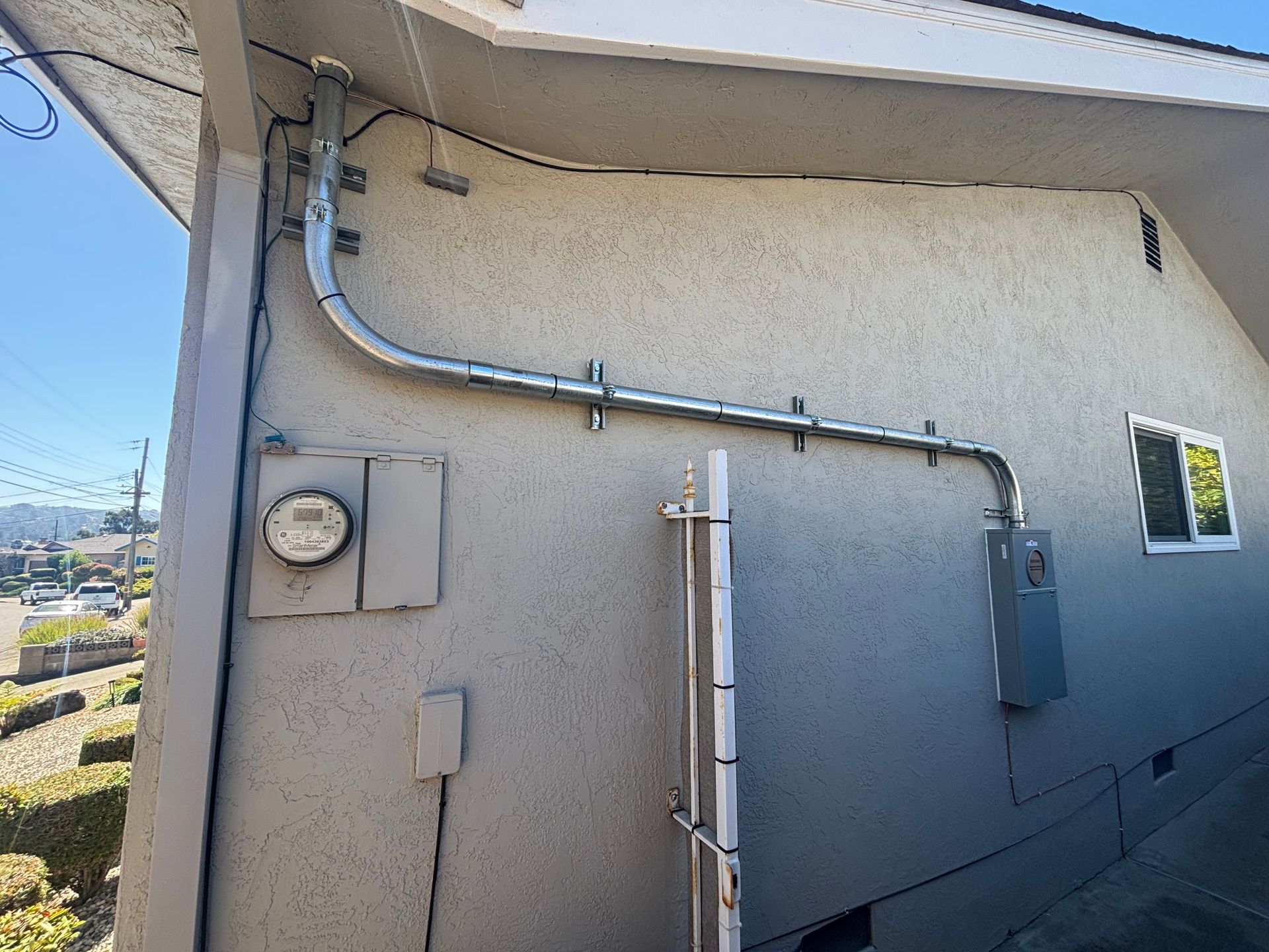 Electrical conduit on an exterior stucco wall of a building; meter box, windows, and sky visible.