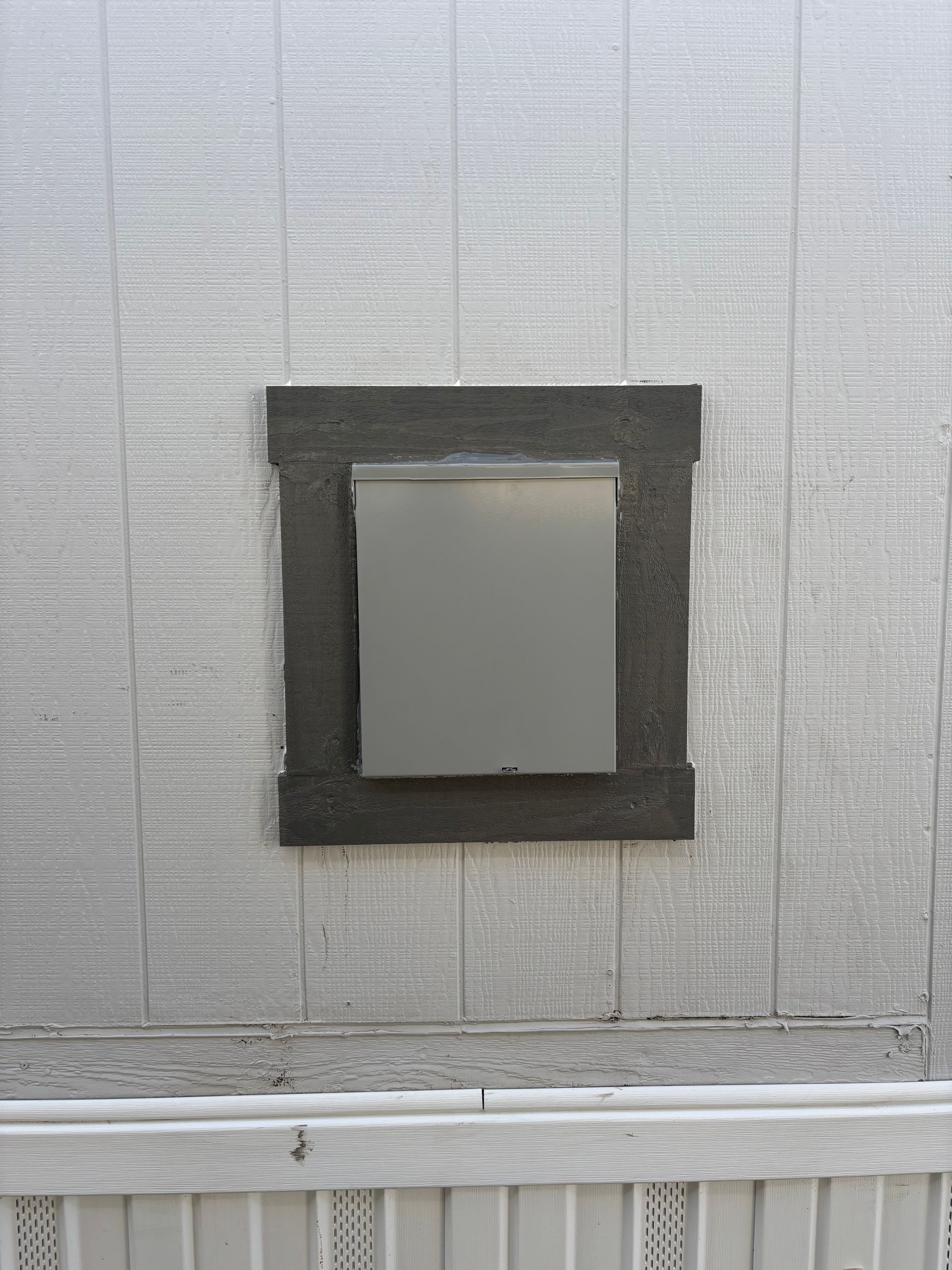 Gray mailbox mounted in a dark square frame on a silver siding wall.