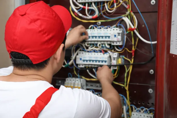 Electrician in red cap and overalls working on wiring in a circuit breaker panel.