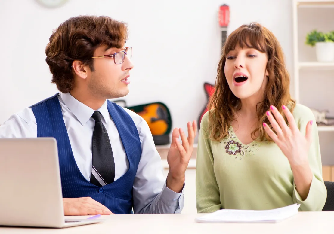 Καθηγητής μουσικής μαθαίνει σε μια γυναίκα να τραγουδάει. Βρίσκονται σε ένα φωτεινό γραφείο με ανοικτό έναν υπολογιστή.