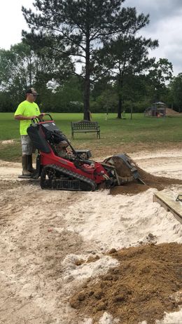 A man in a neon shirt operates a small, red tracked skid steer, moving sand in an outdoor setting.