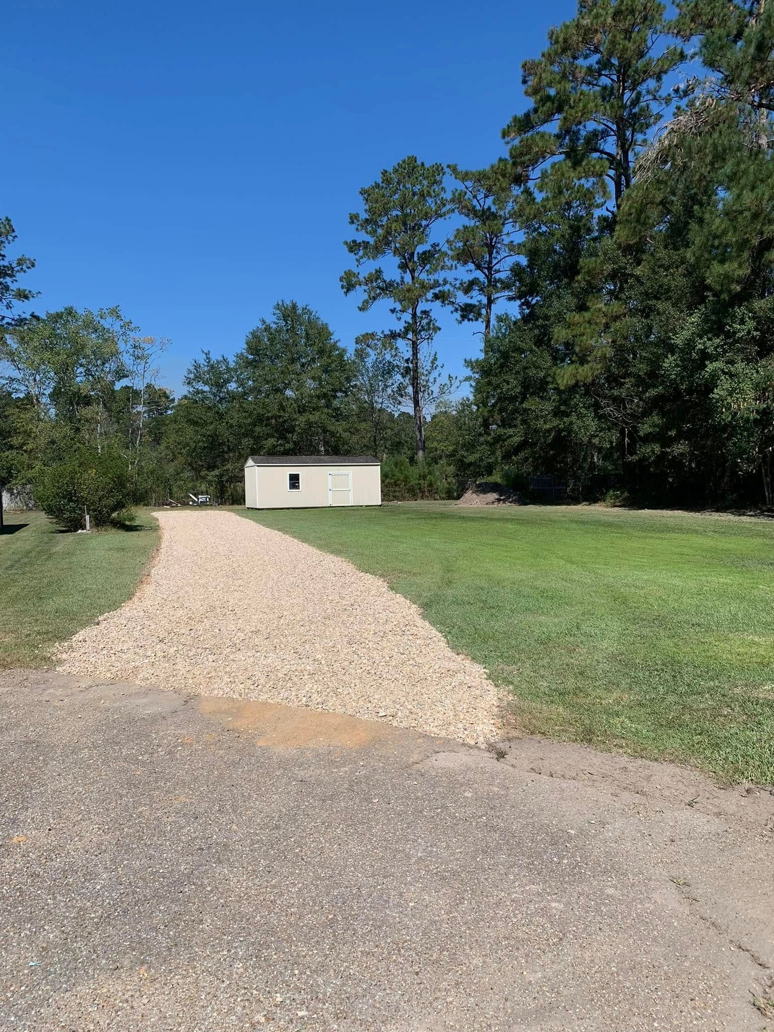 Gravel driveway leading to a shed in a grassy area, trees in the background under a clear blue sky.