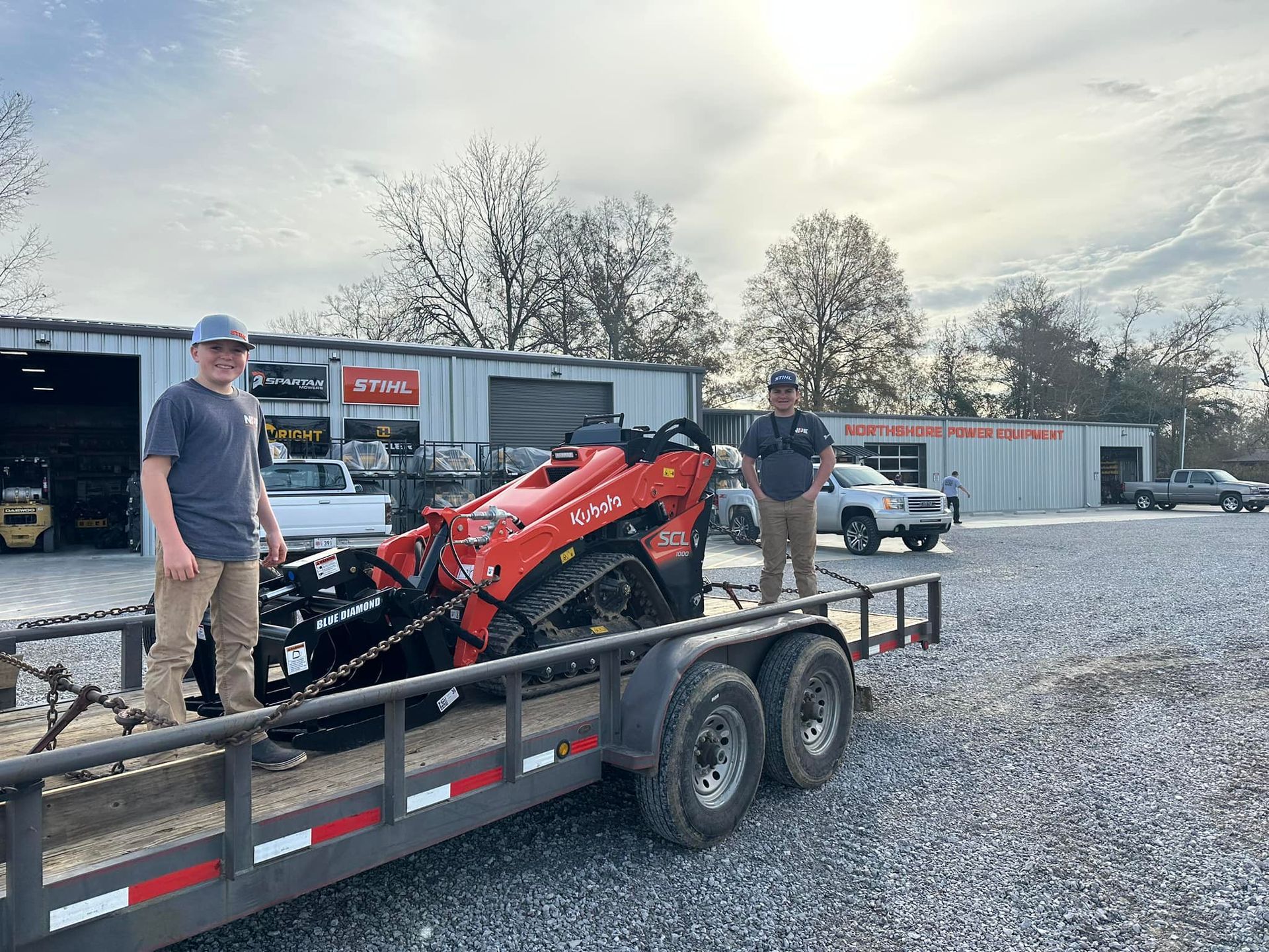 Two men stand on a trailer with a red mini skid steer, parked outside a business. One man faces forward, and the other looks at the camera.