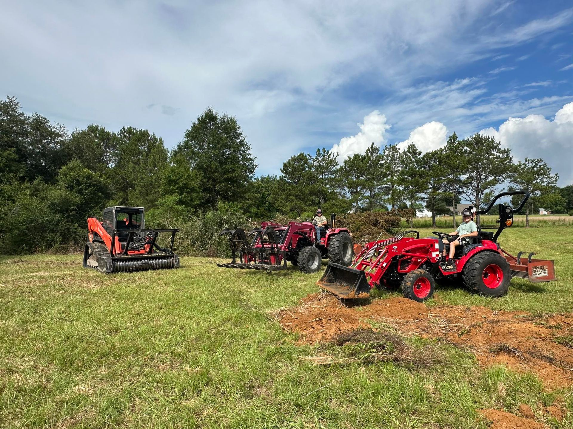 Three red tractors on a grassy field, one with a front-end loader, another with a tiller, and the third with a bush hog attachment; trees in the background.