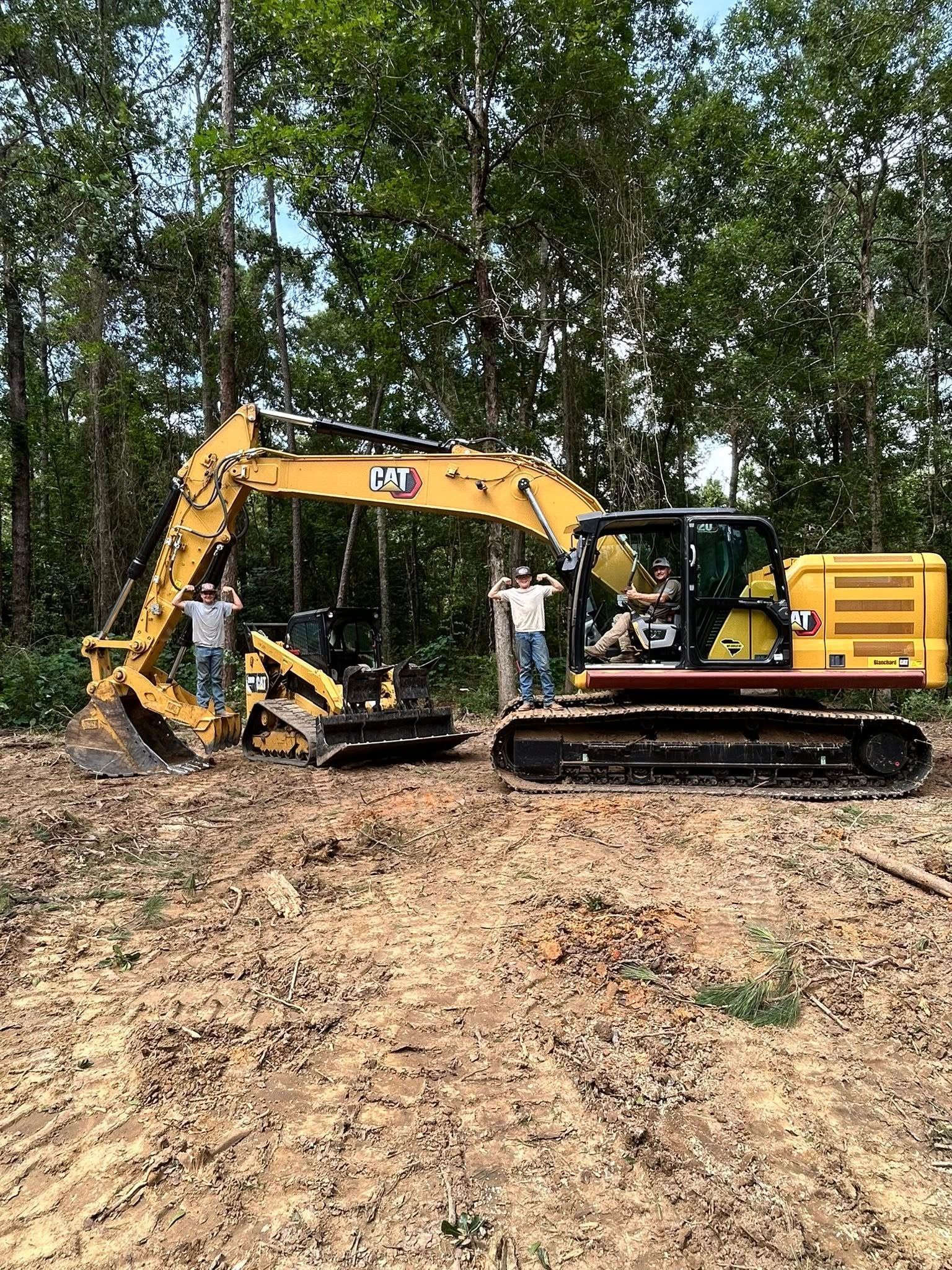 Two men stand near yellow construction equipment, an excavator and a skid steer, in a clearing surrounded by trees.