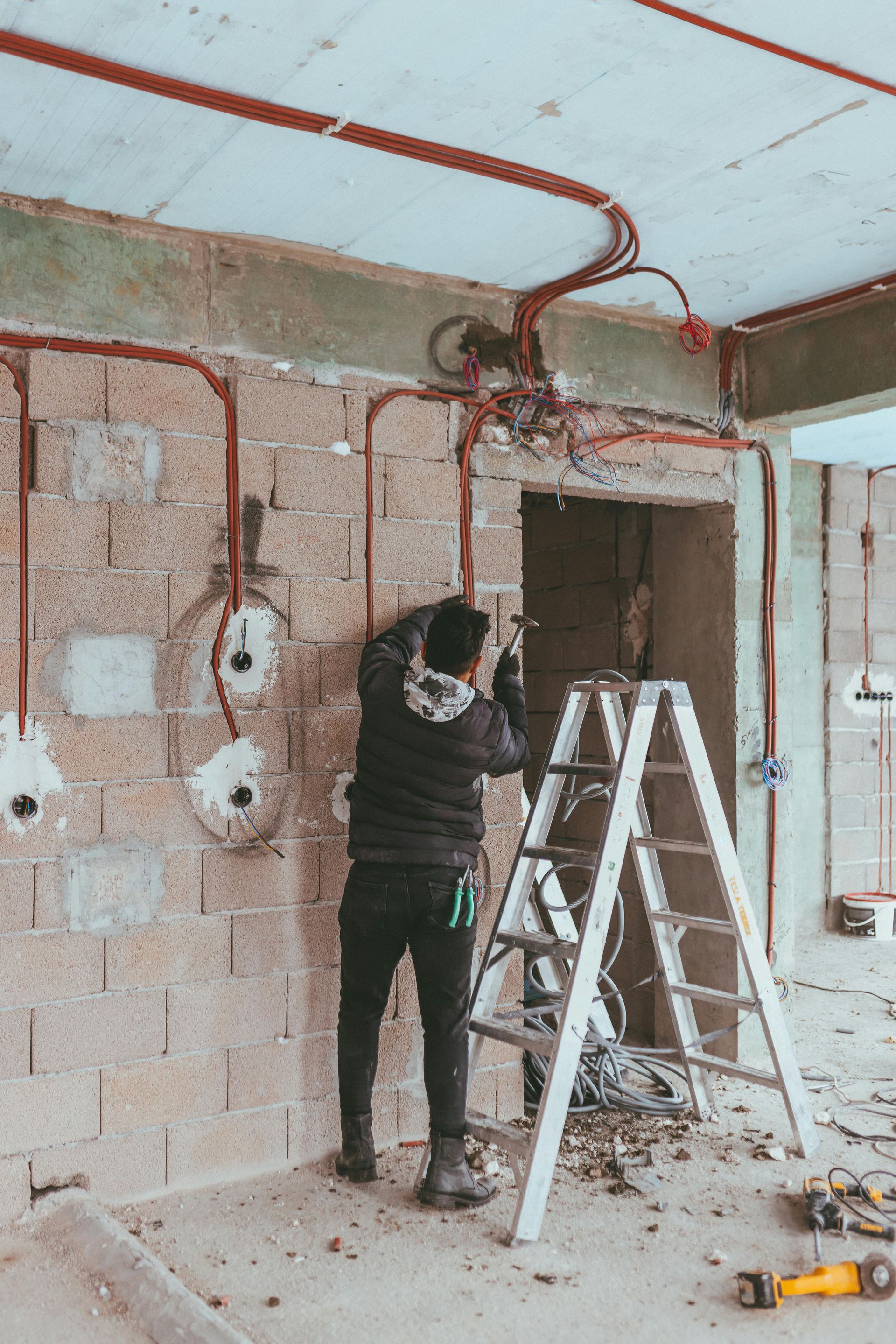 Construction worker on ladder installing electrical wiring in a building. Red conduits run along a brick wall, with outlets visible.