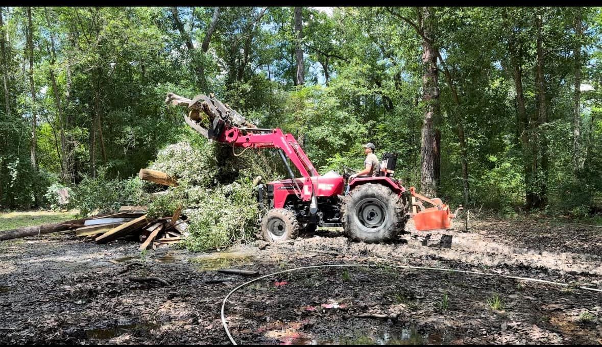 A red tractor with a loader clears debris in a muddy, forested area. A person operates the machinery.