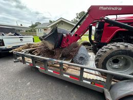 Mahindra tractor loading debris into a trailer, outdoors on a cloudy day. A white pickup truck is visible in the background.