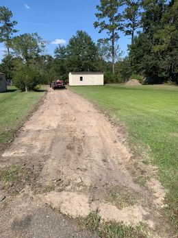 Dirt driveway leading to a small building and trees. A small red tractor is on the driveway. Green grass surrounds the driveway.