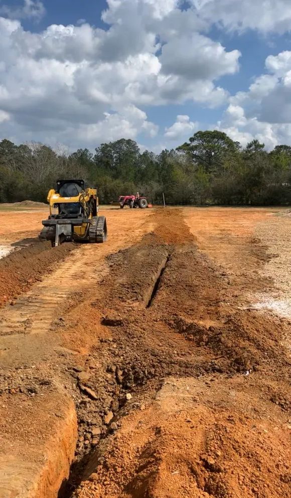 Construction site with a yellow excavator and red vehicle on a dirt field, digging trenches under a partly cloudy sky.