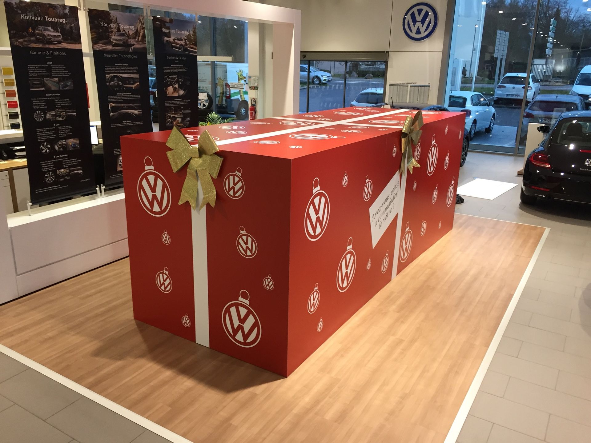 Un grand coffret cadeau rouge est posé sur un plancher en bois chez un concessionnaire automobile.