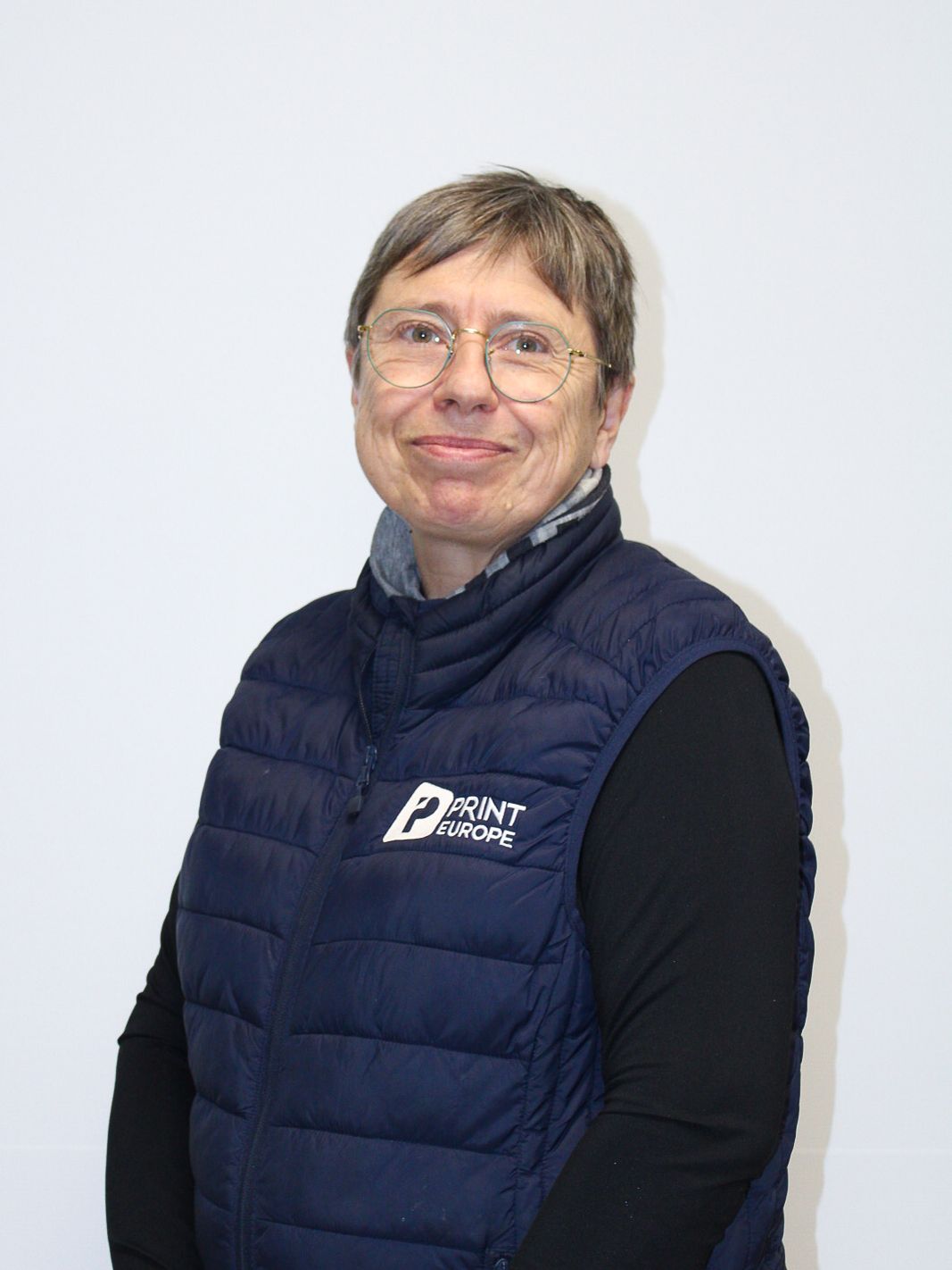 Une femme portant des lunettes et une chemise blanche se tient devant un fond bleu.