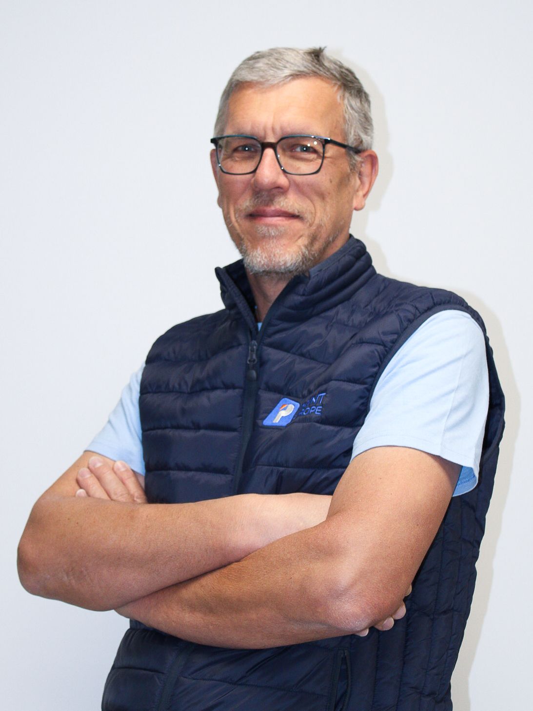 Un homme portant des lunettes et une chemise blanche est debout avec les bras croisés.