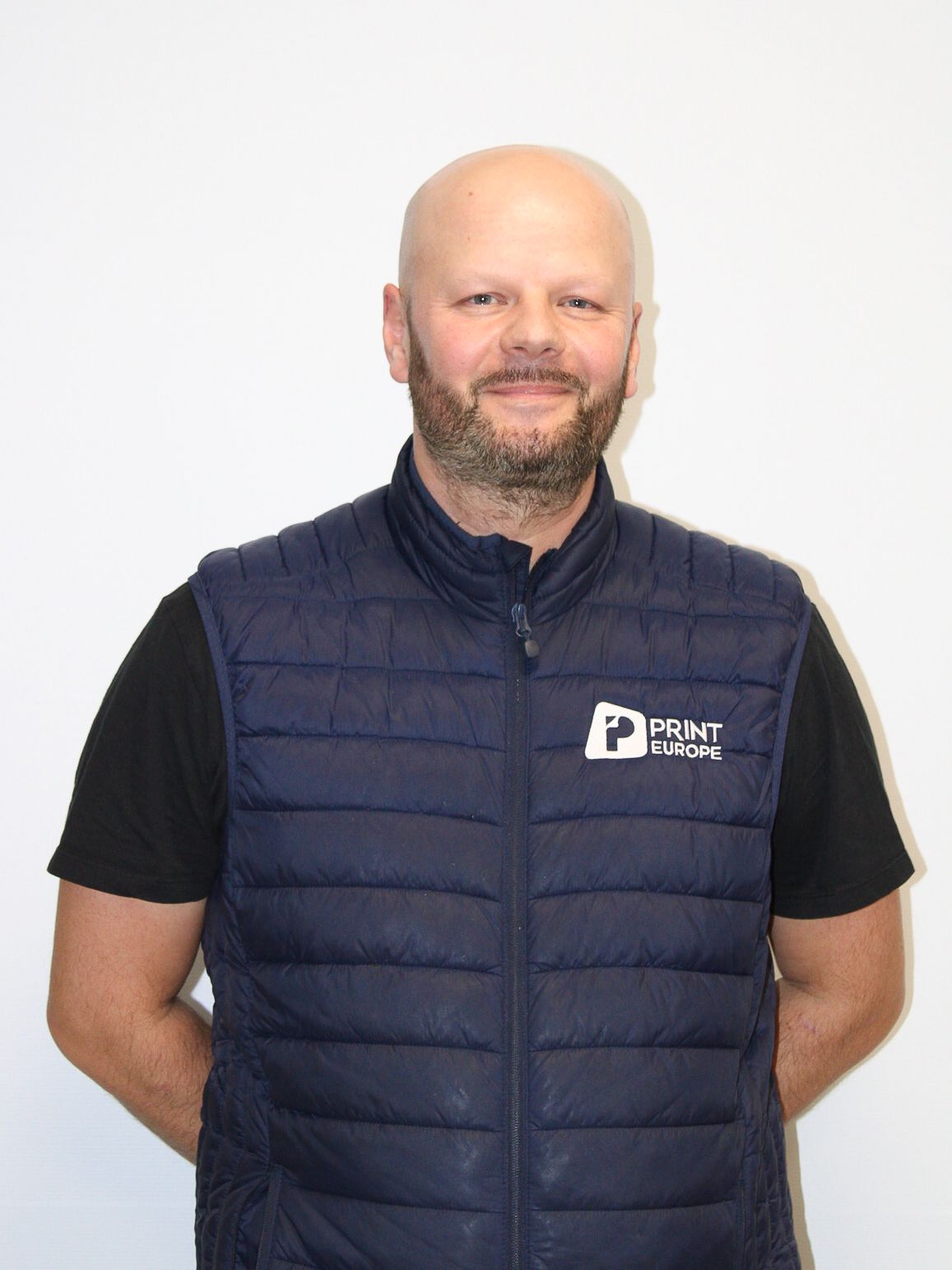 Un homme chauve avec une barbe porte une chemise blanche avec les bras croisés.