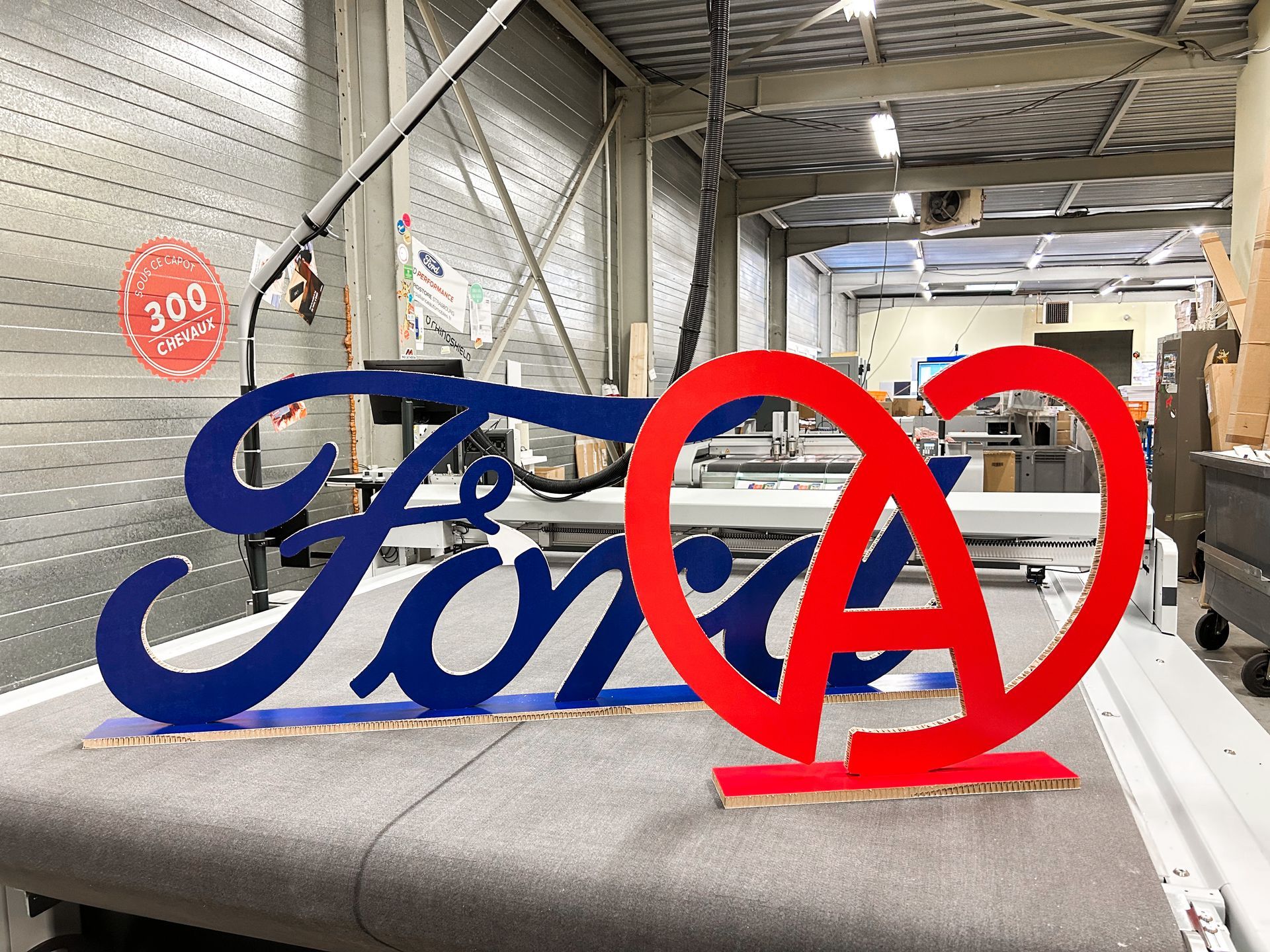 Un grand logo Ford bleu et rouge est posé sur une table dans une usine.