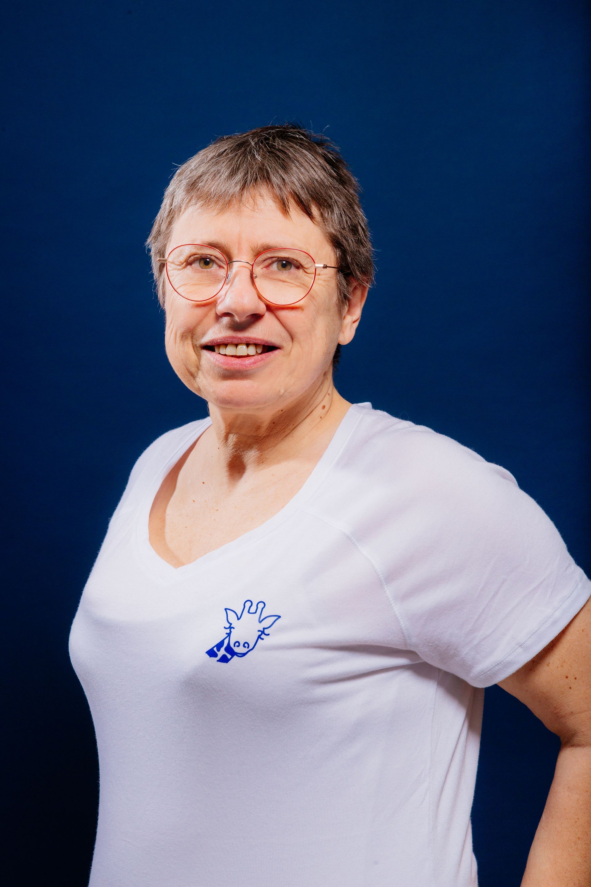 Une femme portant des lunettes et une chemise blanche se tient devant un fond bleu.