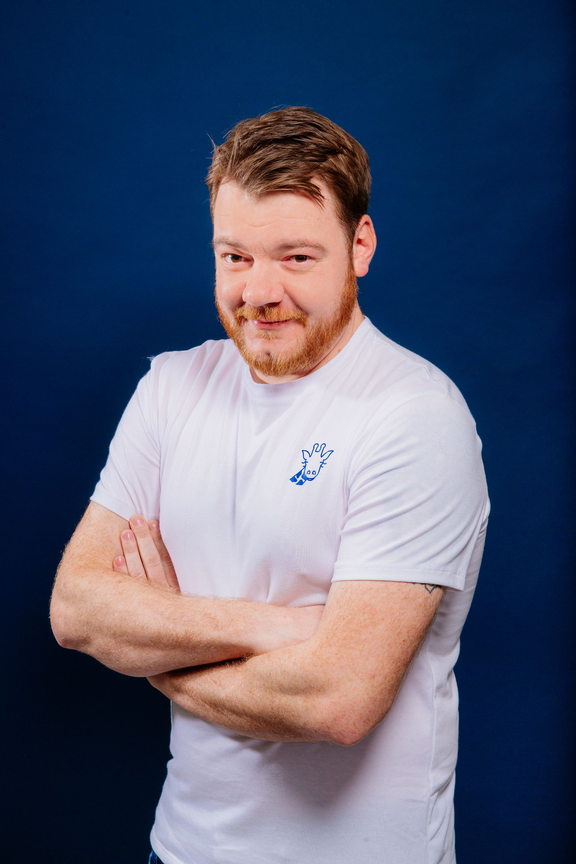 Un homme avec une barbe porte un t-shirt blanc avec les bras croisés.