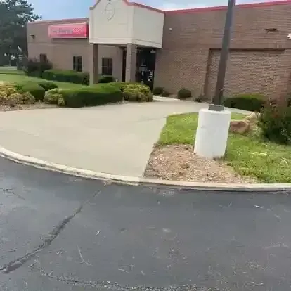 The exterior of a red-brick commercial building with a beige entrance, a concrete walkway, and landscaped shrubs.